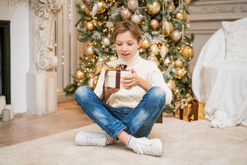 Cute boy in sweater sits by the christmas tree with gifts. Caucasian teenager at home with festive...