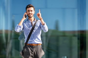 Businessman wearing crossbody bag pointing upward while talking on mobile phone outdoors