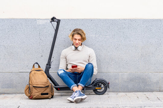 Smiling man with backpack using smart phone while sitting on electric push scooter