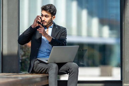 Angry businessman talking on mobile phone while sitting with laptop outdoors