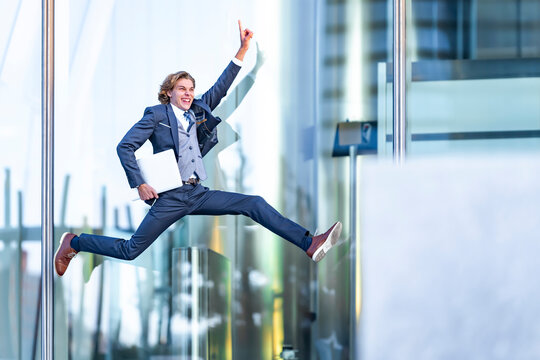 Excited male professional with laptop pointing while jumping against office building