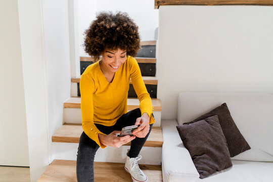 Portrait of smiling young woman sitting on stairs at home looking at cell phone