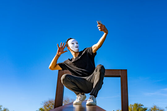 Young man wearing white mask waving hand to video call through mobile phone while crouching on retaining wall