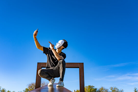 Man with white mask taking selfie through mobile phone while crouching against sky