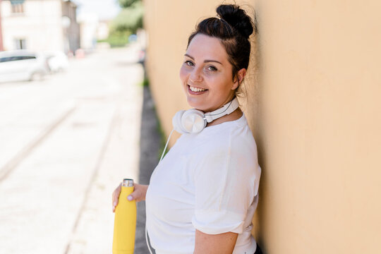 Portrait of a smiling curvy young woman with bottle and headphones leaning against a wall