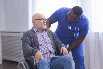Fototapeta premium African-American caregiver and old disabled man in a wheelchair. Professional nurse and handicapped patient in a nursing home. Assistance, rehabilitation and health care.