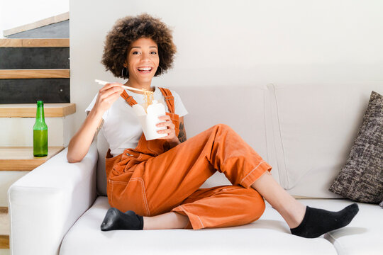 Portrait of happy young woman sitting on the couch at home eating Asian food