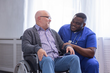 African-American caregiver and old disabled man in a wheelchair. Professional nurse and handicapped patient in a nursing home. Assistance, rehabilitation and health care.