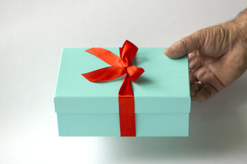The hand of an elderly man holds out a gift in a light green box tied with a red ribbon on a white background