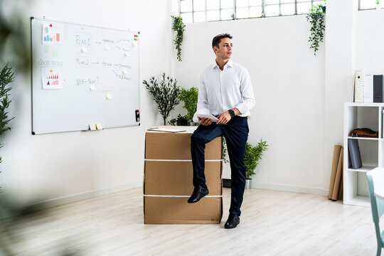 Thoughtful male professional holding clipboard while sitting on cardboard box in office