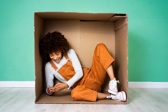 Smiling woman using mobile phone while sitting in cardboard box at new home