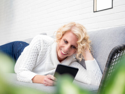 Smiling mature woman lying on couch at home reading e book