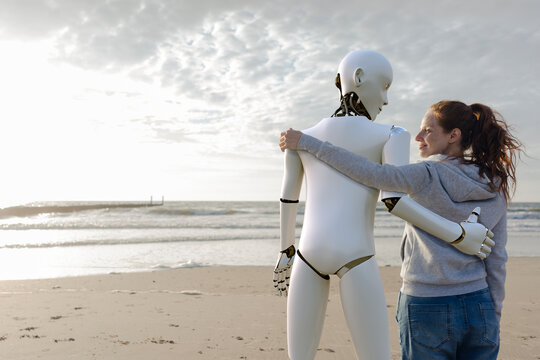 Smiling woman standing arm in arm with robot on the beach