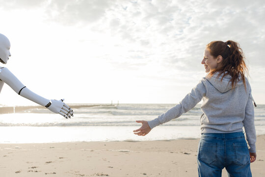Smiling woman standing on the beach reaching for robot hand