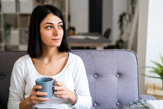 Thoughtful young woman with coffee cup in living room looking away