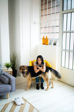 Smiling young woman leaning on dog while sitting on armchair in living room