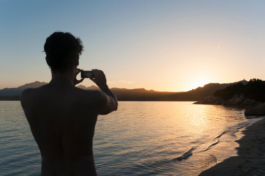 back view of man taking photo of sunset with cell phone, Sardinia, Italy