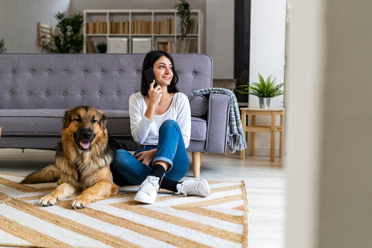 Smiling woman talking on phone while sitting with dog on carpet at home