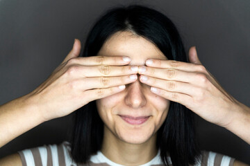 Smiling young woman covering eyes with hands against gray background