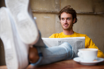 Portrait of young man with newspaper relaxing with feet up in a coffee shop