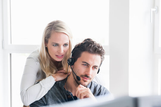 Businesswoman Leaning On Male Customer Service Representative Wearing Headset While Using Computer In Office