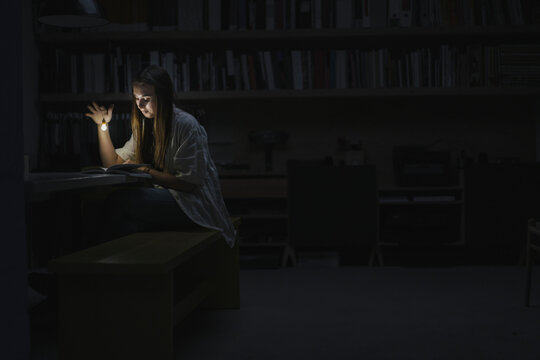 Young woman sitting in office, reading book at night with a torch