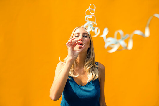Beautiful woman blowing confetti against yellow wall on sunny day