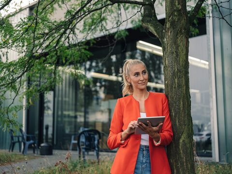 Businesswoman with digital tablet looking away while standing by tree outside office building