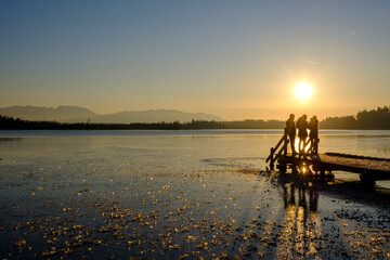 Silhouettes of three people standing on jetty at frozen Kirchsee by sunset, Bavaria, Germany