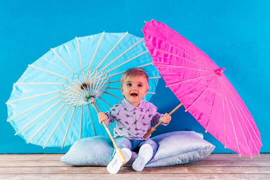 Playful Baby Boy Playing With Paper Umbrellas While Sitting On Pillow In Front Of Blue Wall