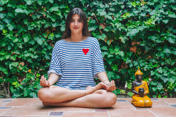 Young woman meditating by Buddha sculpture in backyard