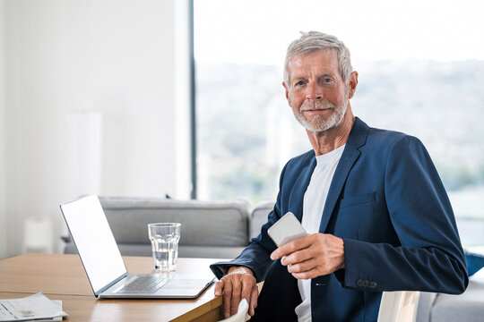 Portrait of senior businessman with laptop and cell phone sitting at desk at home