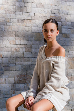 Stylish teenage girl wearing off shoulder dress posing while sitting against brick wall