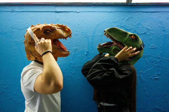 Male and female friends listening to music through headphones while wearing dinosaur mask against blue wall
