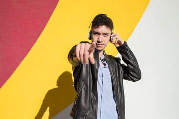 Young man pointing while listening music through headphones against colorful wall on sunny day