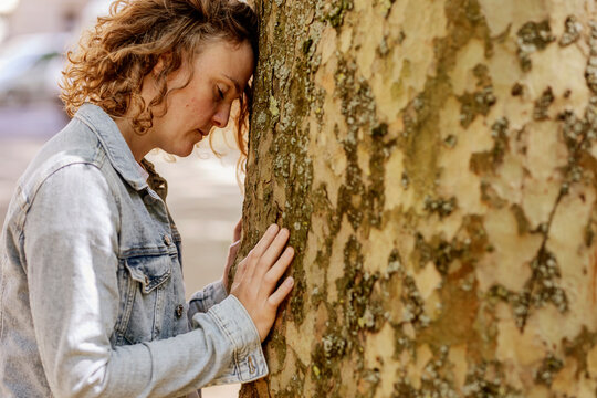 Sad mid adult woman with eyes closed leaning on tree trunk in park
