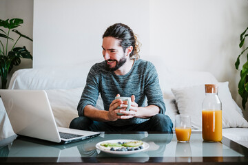 Young man with coffee cup using laptop while having breakfast sitting on sofa at home