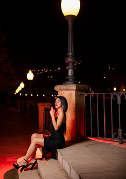 Young woman wearing black evening dress sitting on stairs at night smoking cigarette