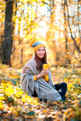 Portrait of a woman in a yellow knitted hat with a cup of hot drink in her hands in the autumn forest. Autumn landscape. Autumn mood
