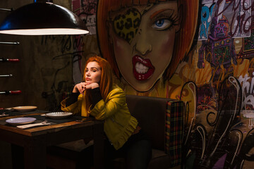 Portrait of redheaded young woman waiting in a pub in front of graffiti wall