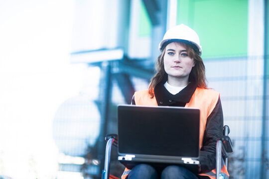 Portrait of young technician with safety helmet and vest in wheelchair working on laptop outdoors
