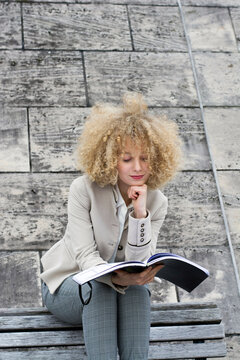 Portrait of blond woman with ringlets sitting on bench looking at booklet