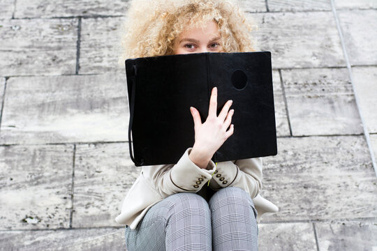 Blond woman with ringlets hiding behind black folder