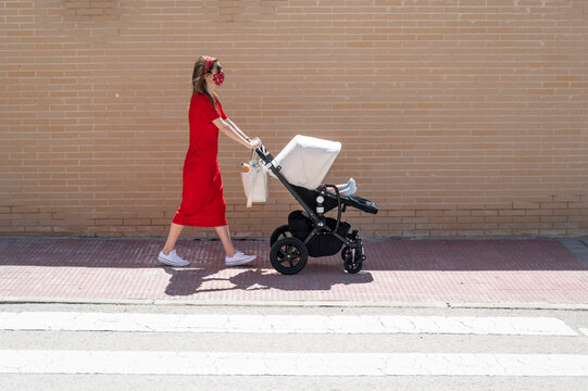 Mother wearing mask pushing son in baby carriage while walking on sidewalk