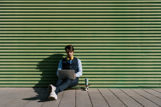 Businessman using laptop while sitting on skateboard against green wall