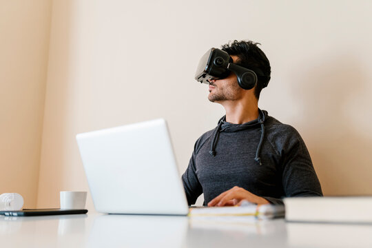 Young man sitting with laptop wearing virtual reality simulator in living room