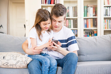 Teenage boy sitting with his little sister on the couch at home looking at cell phone