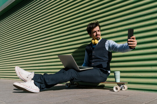 Young businessman with laptop taking selfie while sitting on skateboard against wall