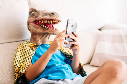 Boy wearing dinosaur mask and cape using smart phone while sitting on sofa in living room