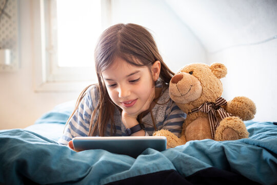 Portrait of smiling girl lying on bed with teddy bear using digital tablet